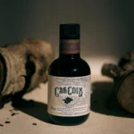 A 250ml bottle of Cas Coix extra virgin olive oil (2025 harvest) standing between rustic wooden logs, produced by Peter Lehner at the Blacknose Ibiza estate.