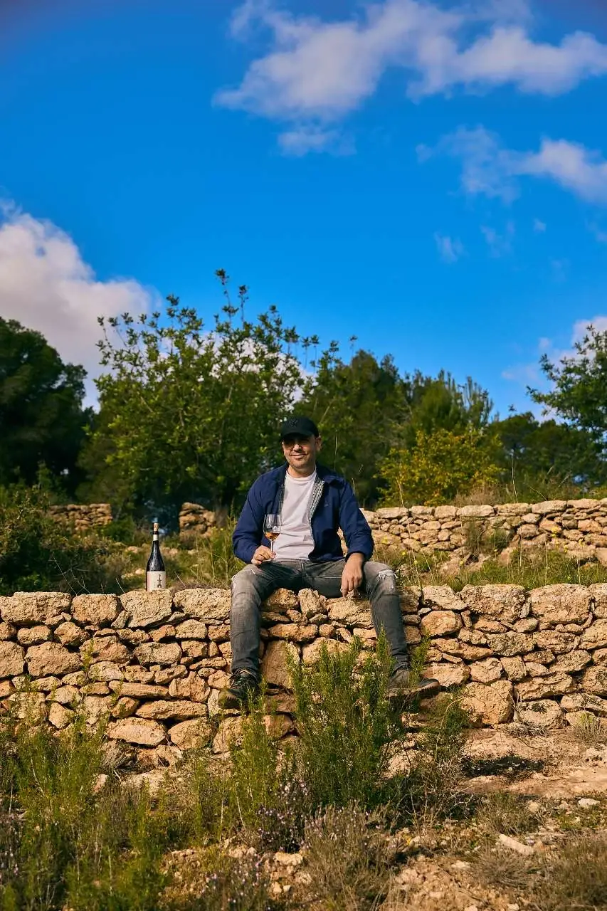 David Lorenzo Álvarez, technical director and viticultor at Ibizkus winery, sitting on a traditional dry-stone wall with a glass of rosé wine; David introduced Peter Lehner to Toni Xumeu de Can Vinya to save his Ibiza wine production.